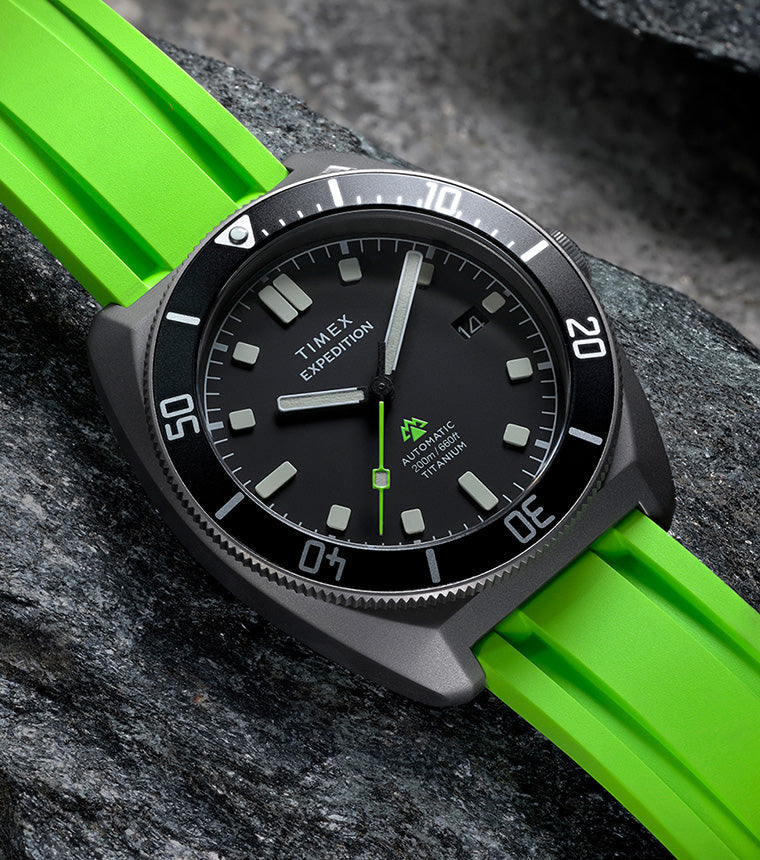 Expedition® Pioneer Titanium Automatic resting on rugged rocks, highlighting the black dial, rotating bezel, and vivid green strap.