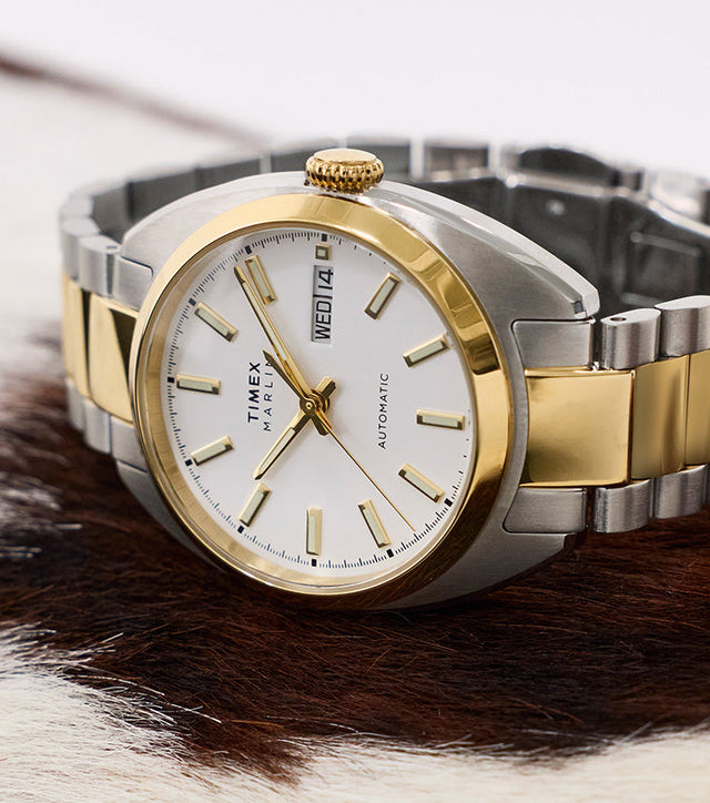Timex Marlin® Draper Automatic two-tone stainless steel watch resting on a textured surface, featuring a white dial and gold-tone accents.