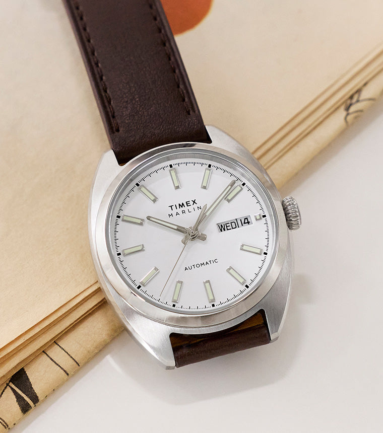 Timex Marlin® Draper Automatic watch with a brown leather strap resting on an open book, featuring a white dial and stainless steel case.