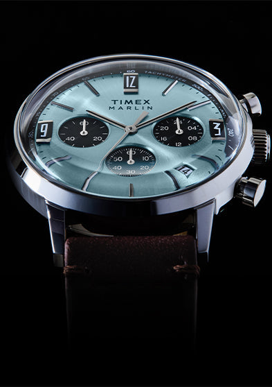 Timex Marlin Chronograph Tachymeter watch with a light blue dial, black chronograph subdials, tachymeter scale, and brown leather strap, shown at an angled close-up against a black background.