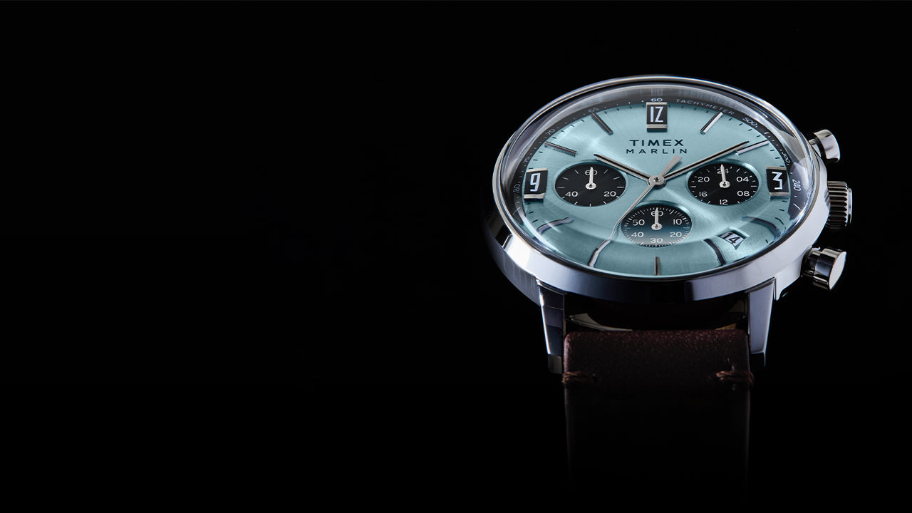 Timex Marlin Chronograph Tachymeter watch with a light blue dial, black chronograph subdials, tachymeter scale, and brown leather strap, shown at an angled close-up against a black background.