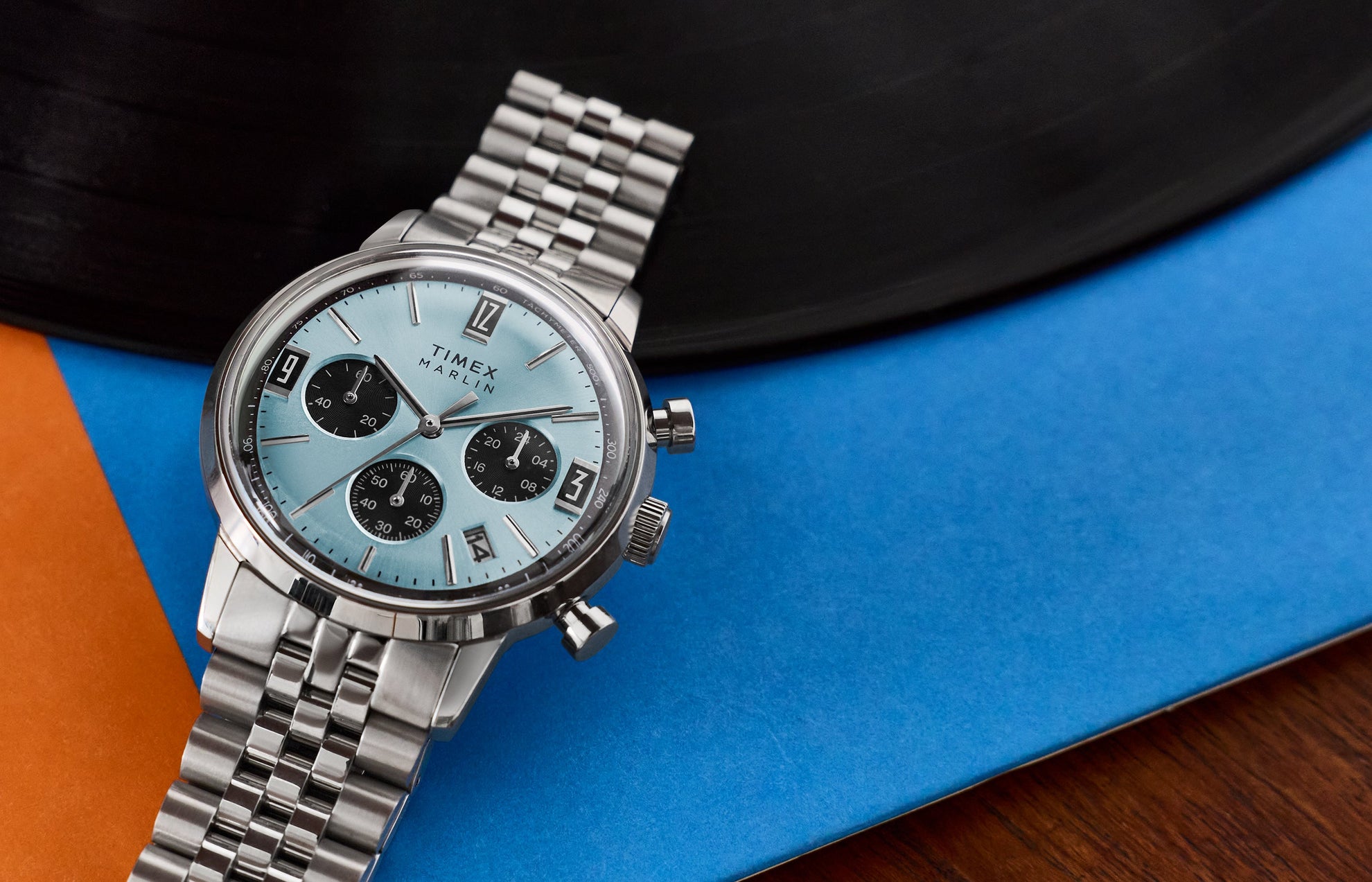 Timex Marlin Chronograph Tachymeter watch with a light blue dial, black chronograph subdials, and stainless steel bracelet, resting on a blue and orange graphic surface.