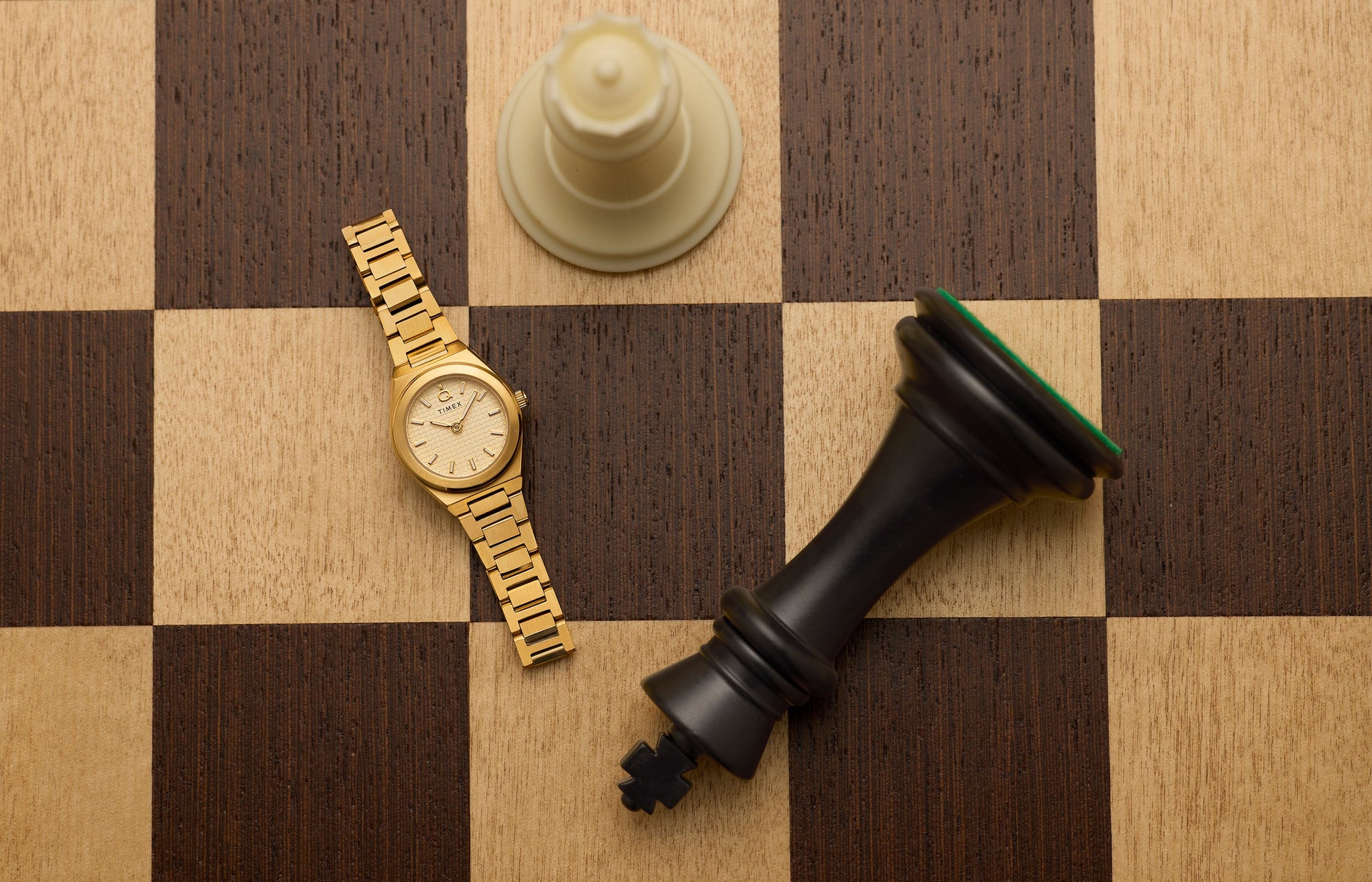 Q Timex® Continental Mini gold-tone watch with textured champagne dial and gold stainless steel bracelet displayed on a wooden chessboard beside oversized chess pieces.