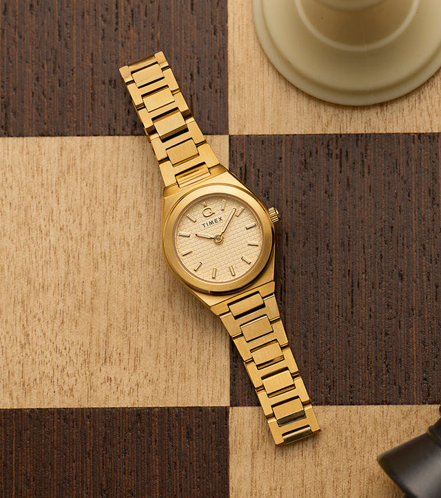 Close-up of Q Timex® Continental Mini in gold-tone stainless steel featuring a champagne textured dial and slim integrated bracelet on a chessboard backdrop.