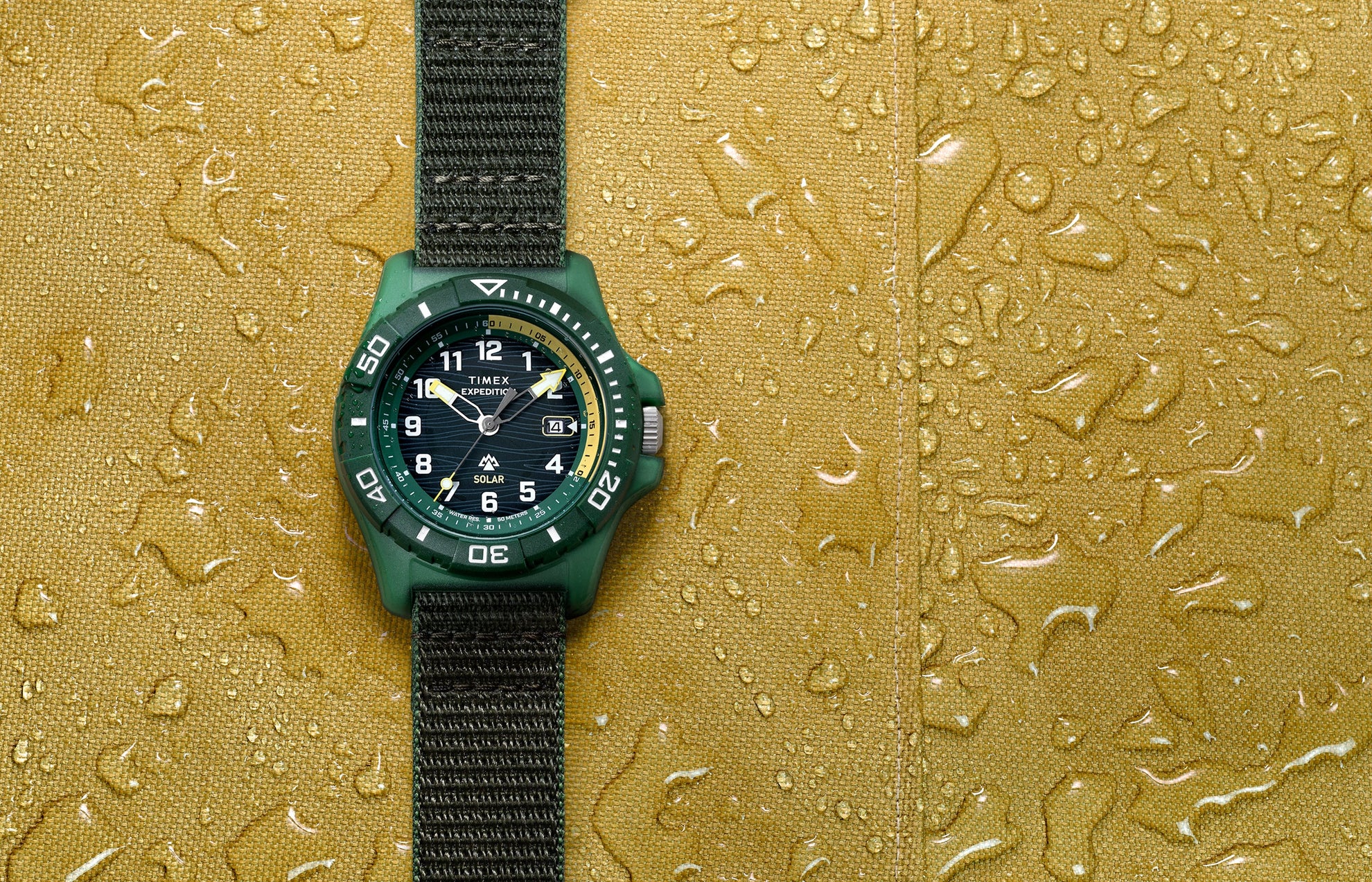Timex Expedition® Freedive Solar watch with green resin case and black fabric strap displayed on yellow water-resistant surface with water droplets.