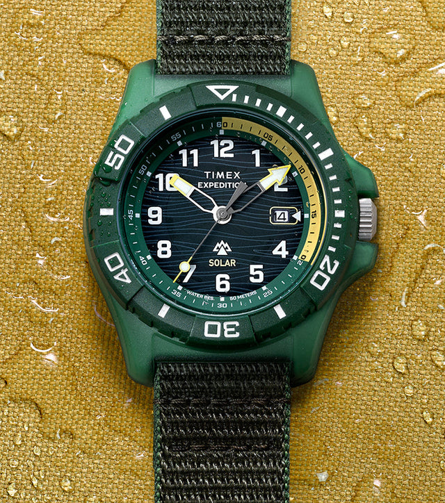 Timex Expedition® Freedive Solar watch with green resin case and black fabric strap displayed on yellow water-resistant surface with water droplets.