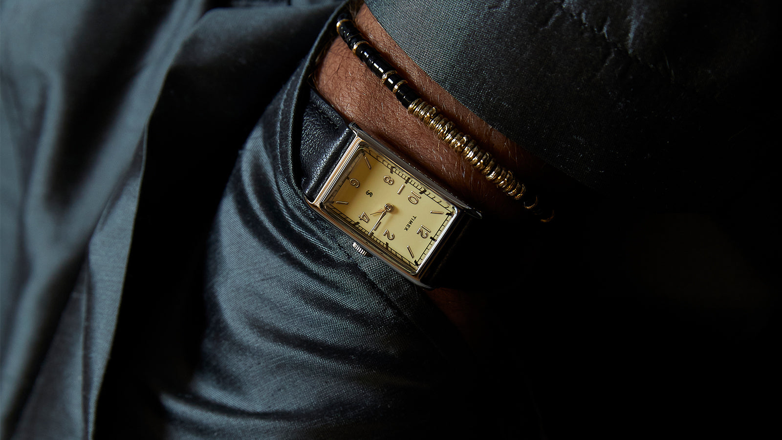 Angled close-up of the Timex x Todd Snyder 1976 Lexington watch with a cream dial and black leather strap worn with a dark jacket sleeve.