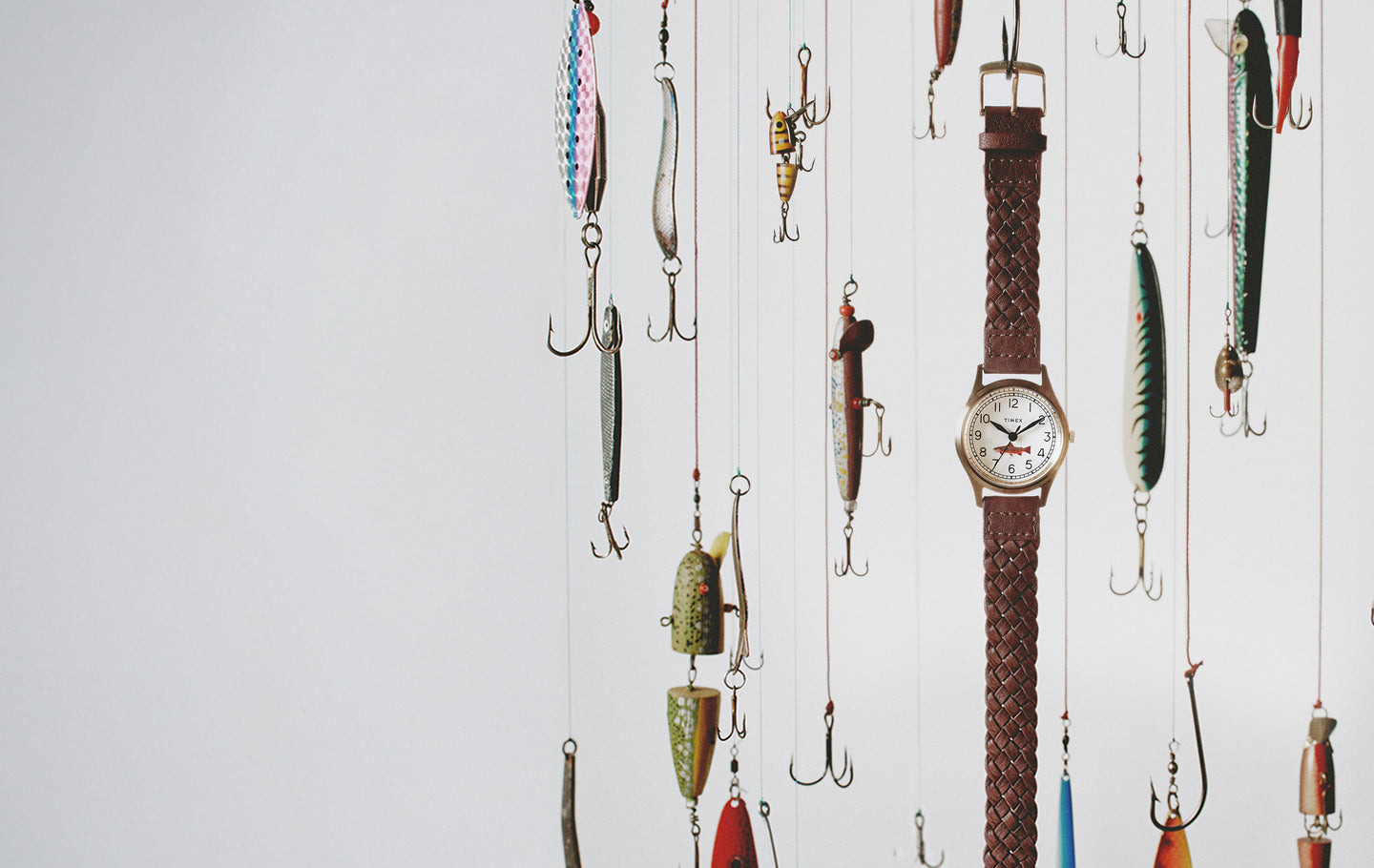 Timex MK1 watch with braided brown leather strap suspended among colorful vintage fishing lures against a soft white background.