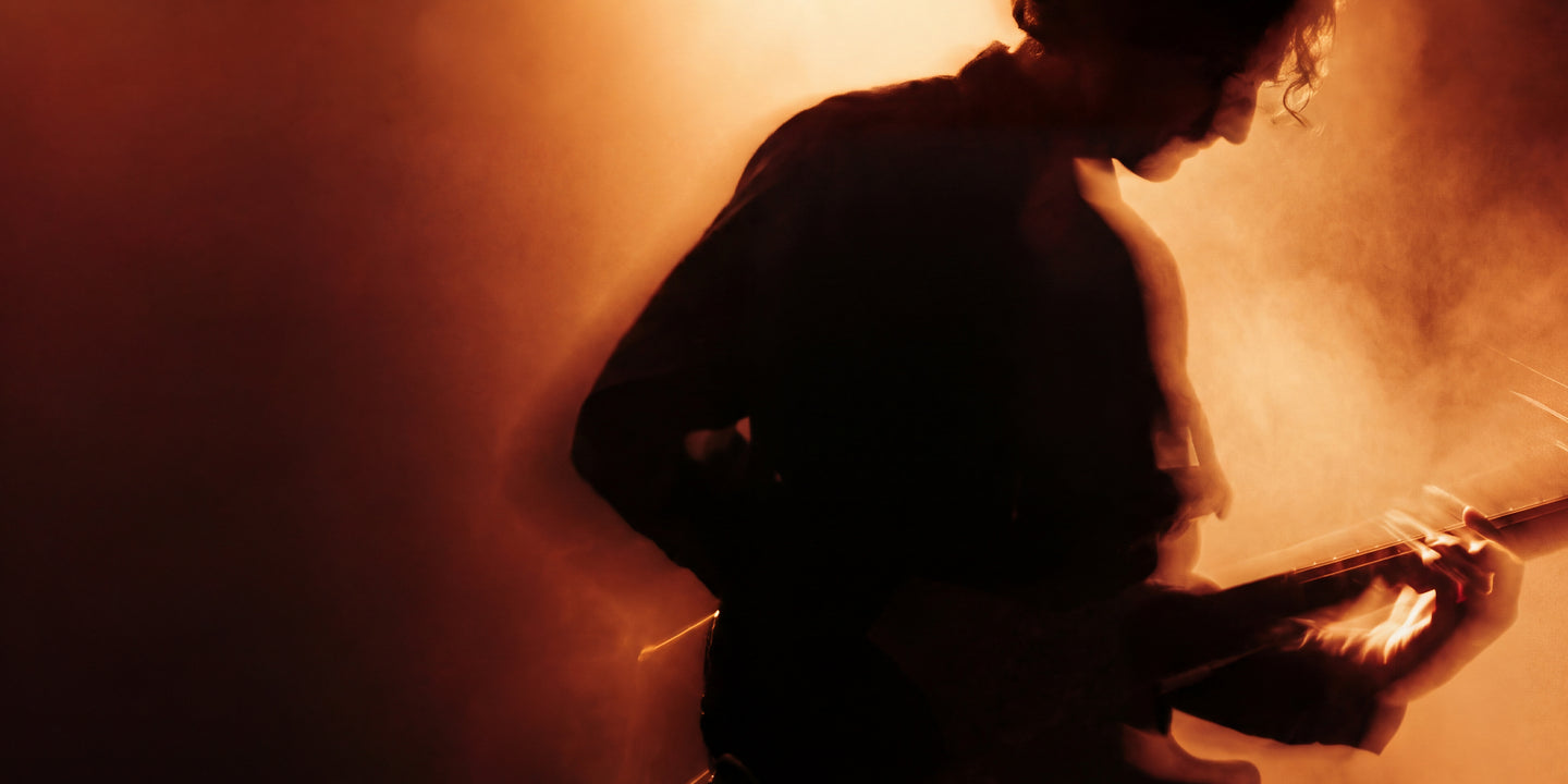 Silhouetted guitarist performing on stage, backlit by warm orange light and haze.