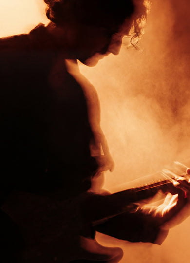 Silhouetted guitarist performing on stage, backlit by warm orange light and haze.