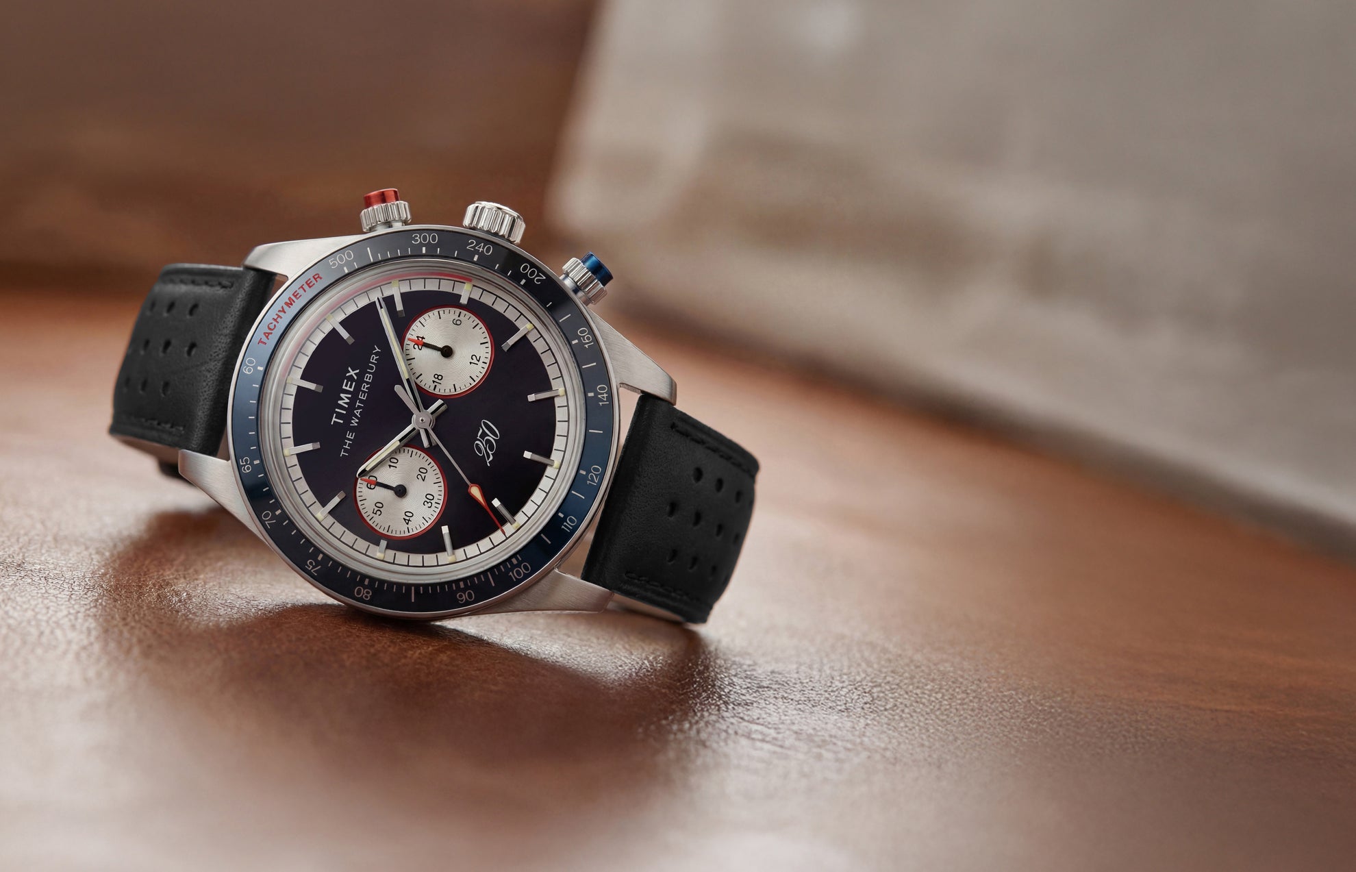 Waterbury Heritage America 250 Chronograph watch with a black leather racing strap, blue tachymeter bezel, and commemorative dial displayed on a wooden surface.