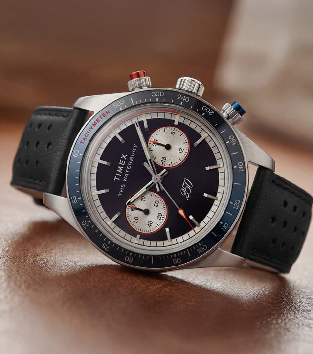 Waterbury Heritage America 250 Chronograph watch with a black leather racing strap, blue tachymeter bezel, and commemorative dial displayed on a wooden surface.