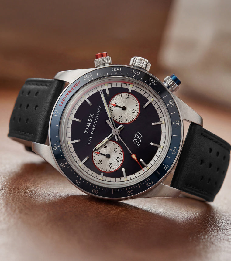 Waterbury Heritage America 250 Chronograph watch with a black leather racing strap, blue tachymeter bezel, and commemorative dial displayed on a wooden surface.