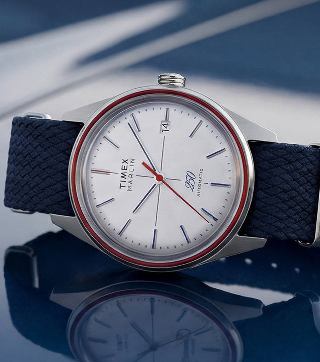 Marlin Jet America 250 automatic watch with a white dial, red bezel accent, red seconds hand, and blue woven strap displayed on a reflective blue surface.