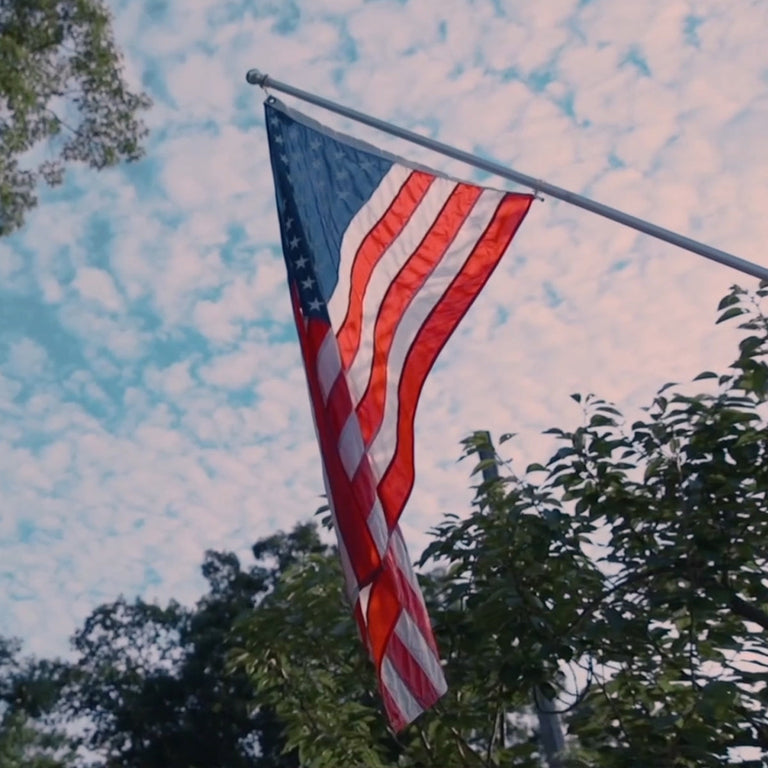American flag waving against a blue sky and trees, symbolizing the patriotic inspiration of the Waterbury America 250 Collection.