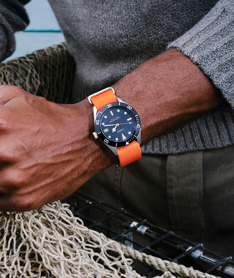 TWG069200 Man wearing a Bespoke Post x Timex Heritage 66 watch with a dark dial and bright orange fabric strap, holding a fishing net near the water.