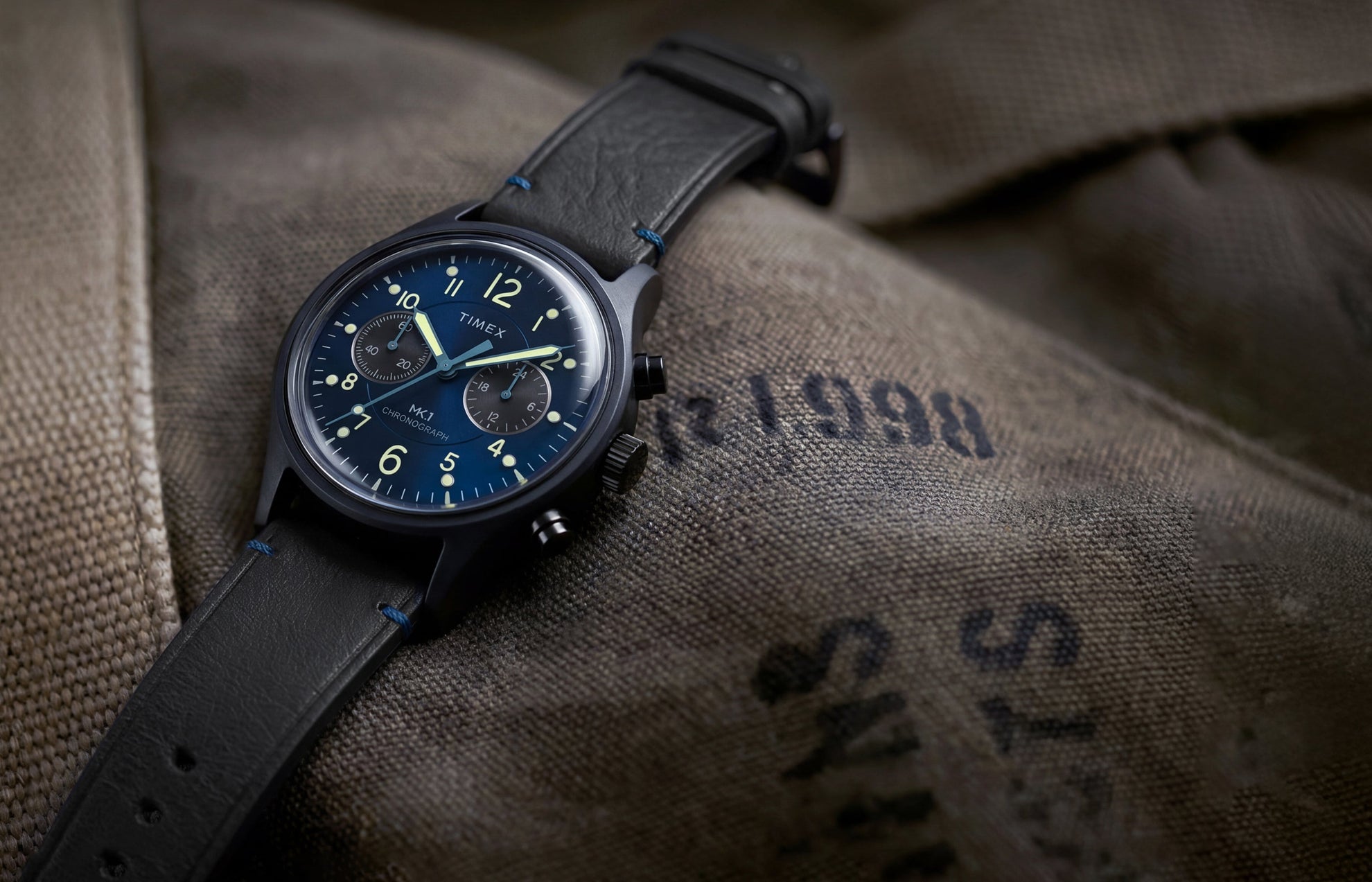 Timex MK1® Chronograph watch with a blue dial, black leather strap, and black case displayed on canvas fabric.