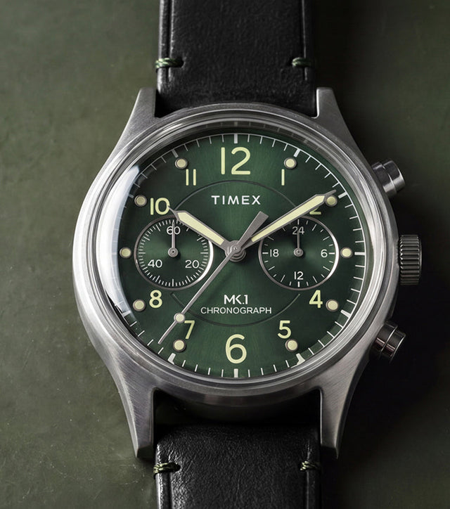 Timex MK1® Chronograph watch with a green dial, black leather strap, and brushed stainless steel case displayed on a dark green surface.