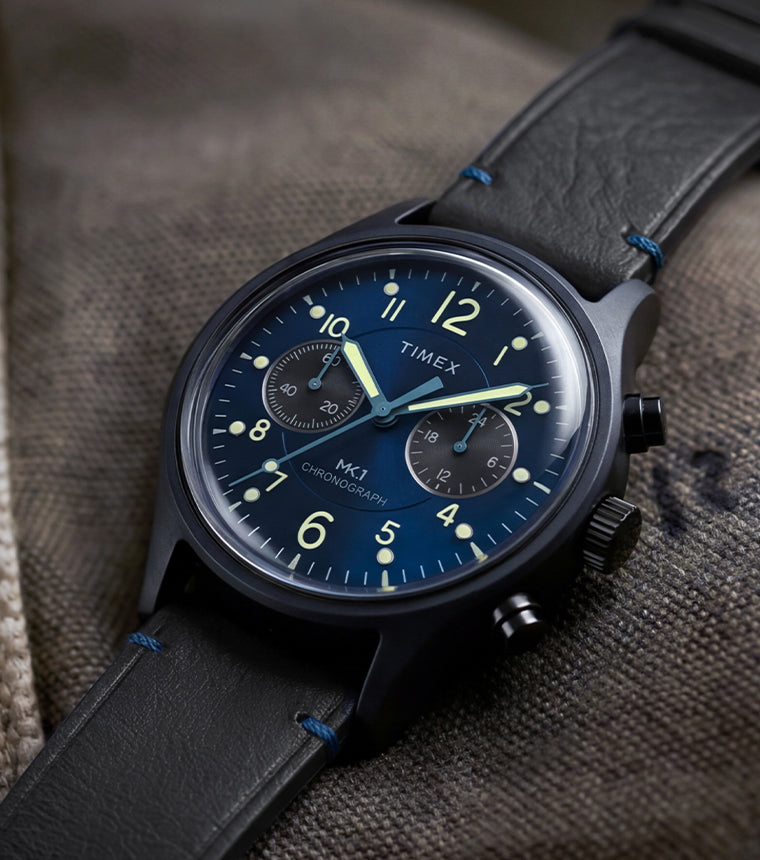 Timex MK1® Chronograph watch with a blue dial, black leather strap, and black case displayed on canvas fabric.