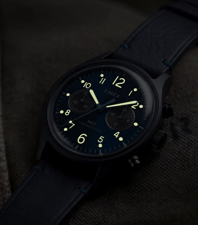 Timex MK1® Chronograph watch showing glowing luminous hands and hour markers on a blue dial in low light.
