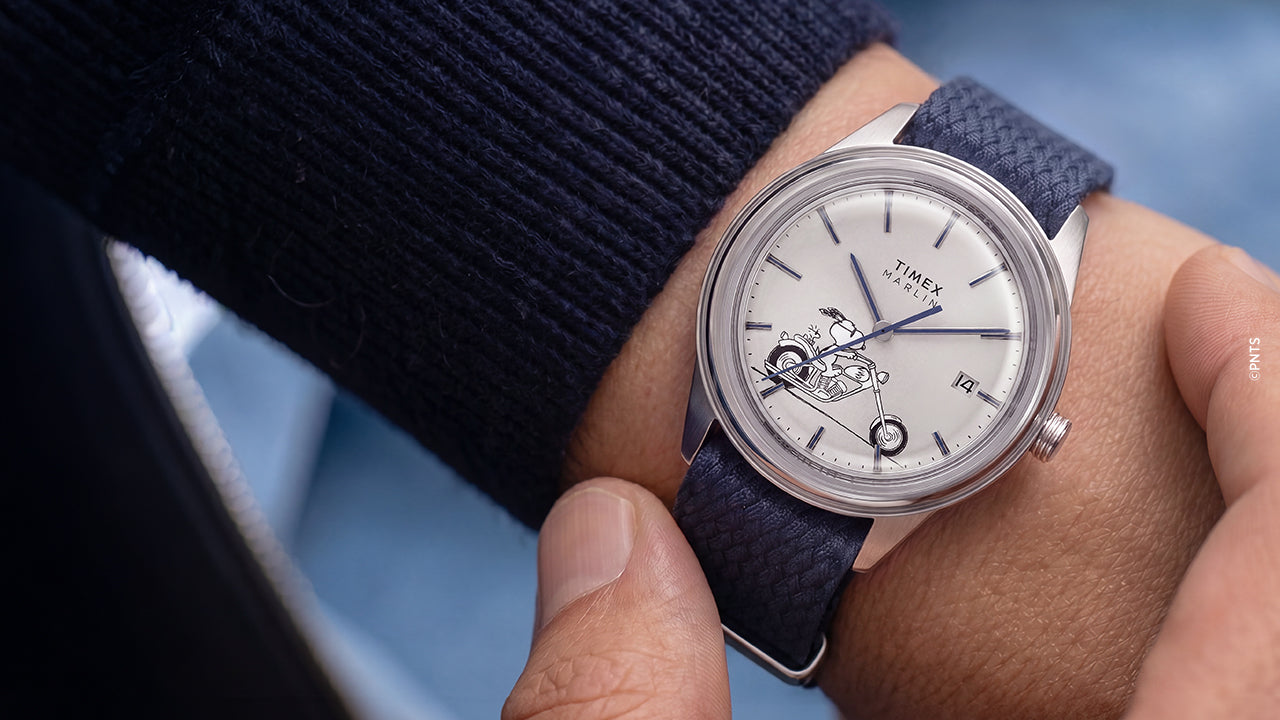 Lifestyle image of the Peanuts® x Timex Marlin® Jet Automatic Easy Rider watch on the wrist, featuring a white dial with Snoopy riding a motorcycle, silver-tone case, and blue woven strap.