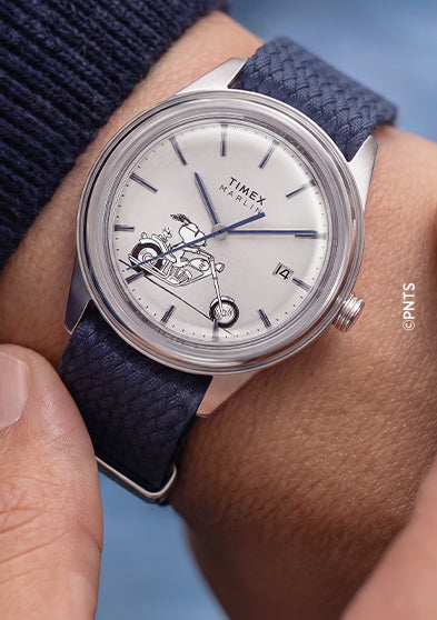 Lifestyle image of the Peanuts® x Timex Marlin® Jet Automatic Easy Rider watch on the wrist, featuring a white dial with Snoopy riding a motorcycle, silver-tone case, and blue woven strap.