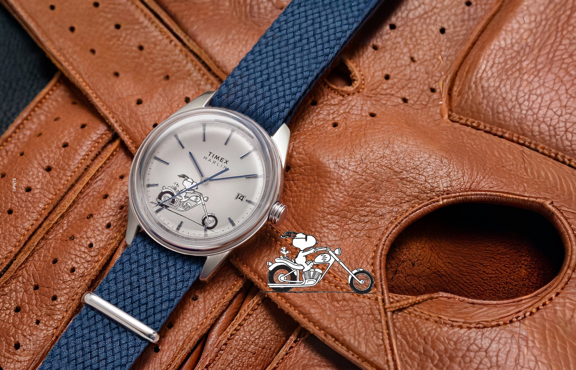 Peanuts® x Timex Marlin® Jet Automatic Easy Rider watch with a silver-tone case, white dial featuring Snoopy riding a motorcycle, and blue woven strap displayed on a brown leather motorcycle jacket.