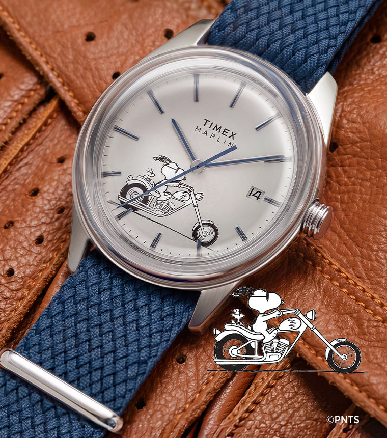 Peanuts® x Timex Marlin® Jet Automatic Easy Rider watch with a silver-tone case, white dial featuring Snoopy riding a motorcycle, and blue woven strap displayed on a brown leather motorcycle jacket.