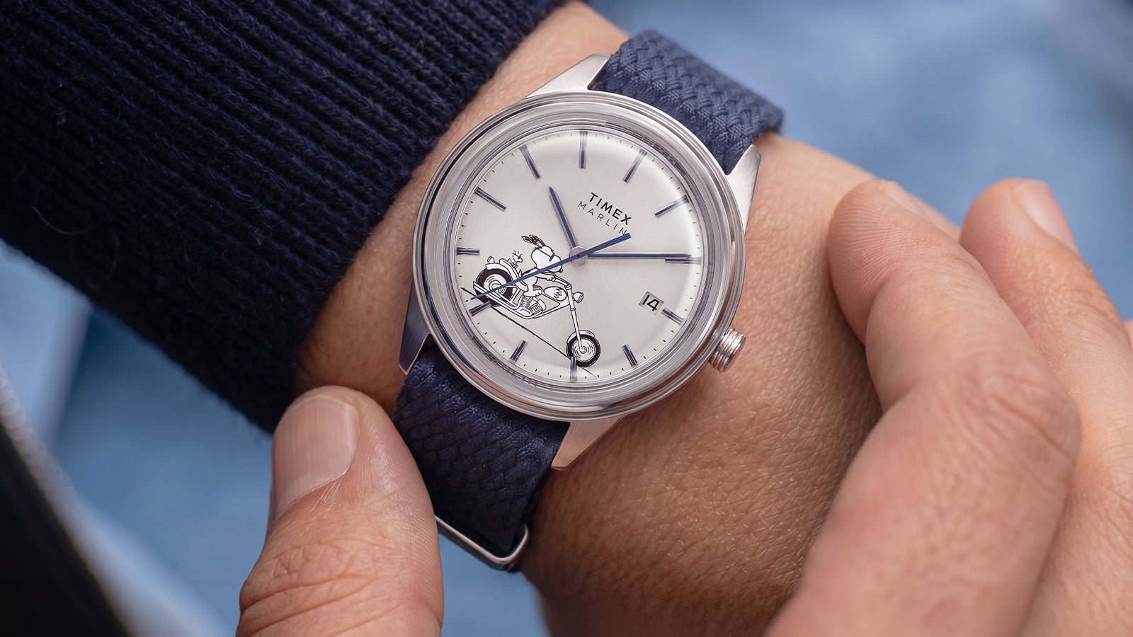 Peanuts® x Timex Marlin® Jet Automatic Easy Rider watch worn on the wrist, featuring a white dial with Snoopy motorcycle artwork, silver-tone case, and textured blue woven strap.