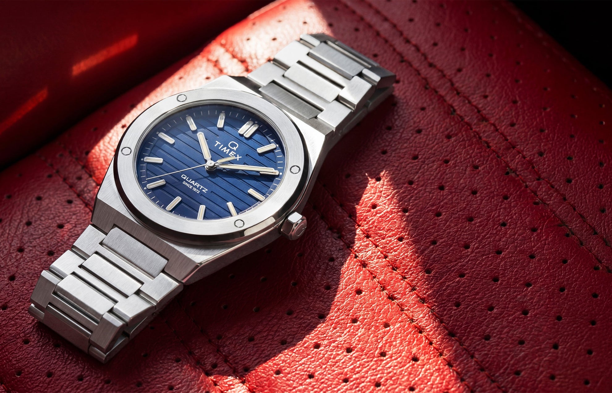 Timex Q Marbella stainless steel watch with a blue textured dial resting on a red perforated leather car seat.