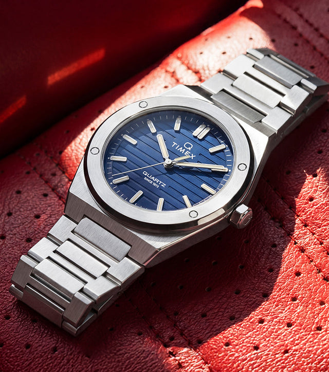 Timex Q Marbella stainless steel watch with a blue textured dial resting on a red perforated leather car seat.