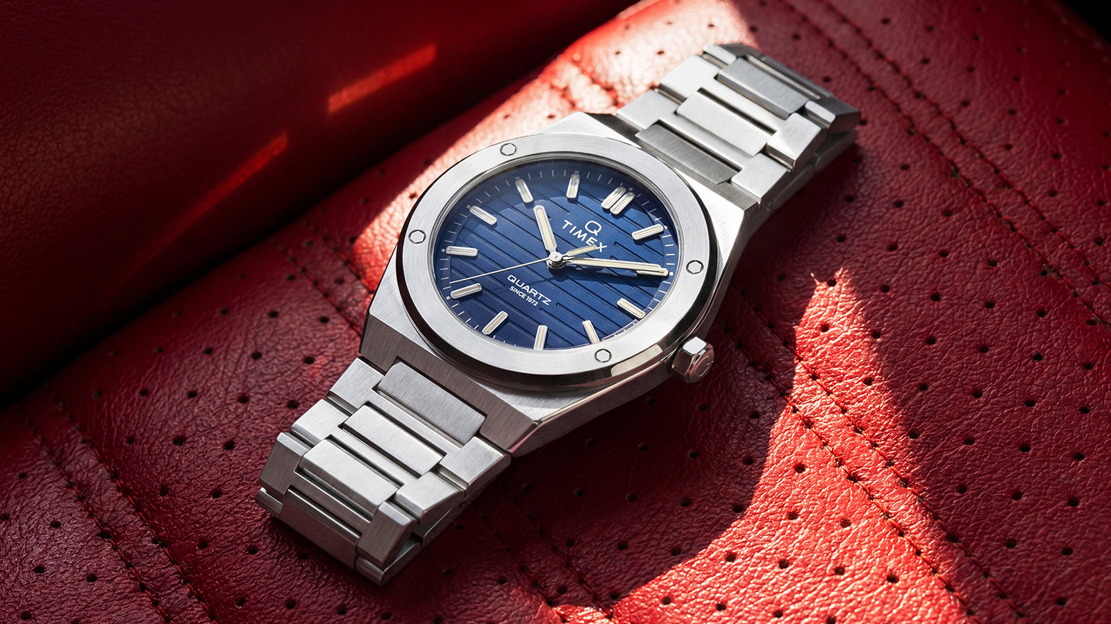 Timex Q Marbella stainless steel watch with a blue dial showcased on a red perforated leather surface.
