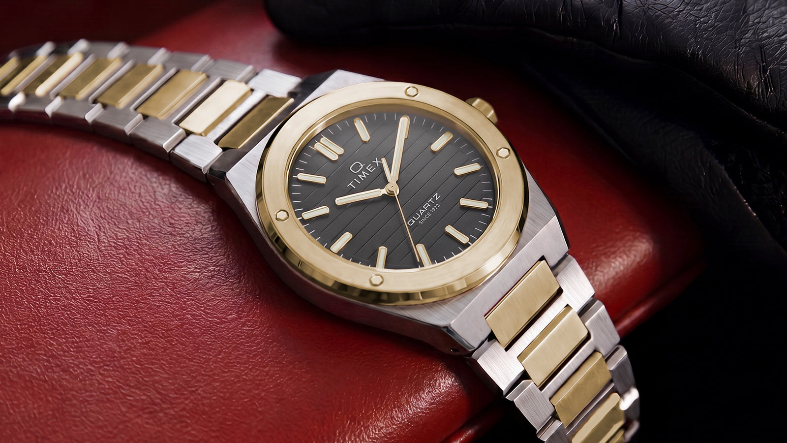 Timex Q Marbella two-tone stainless steel watch with a black textured dial resting on a red leather surface beside a black driving glove.