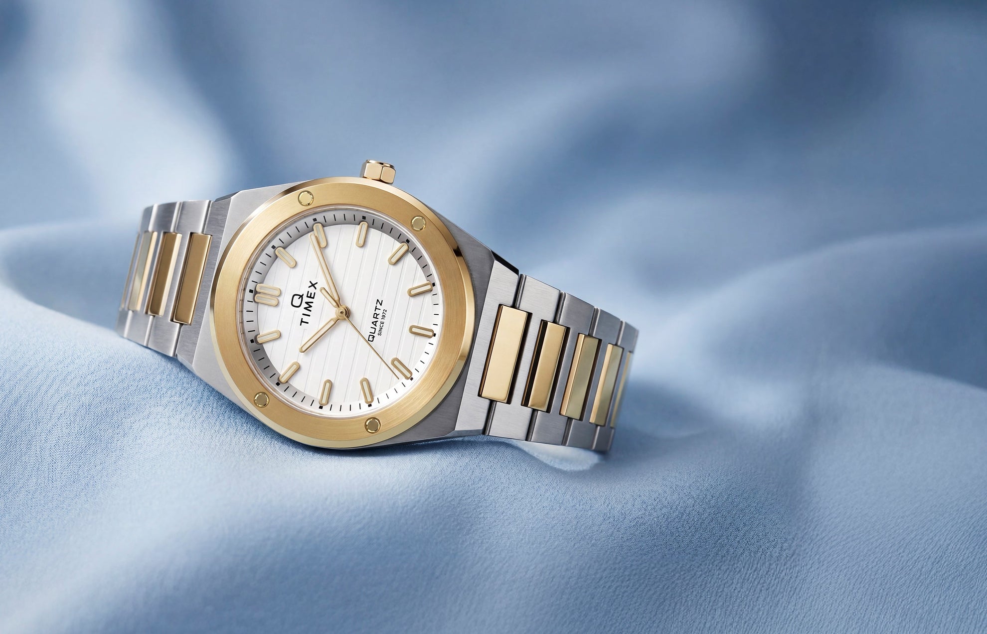 Q Timex Marbella two-tone stainless steel watch with a white dial and gold-tone bezel displayed on soft blue fabric.