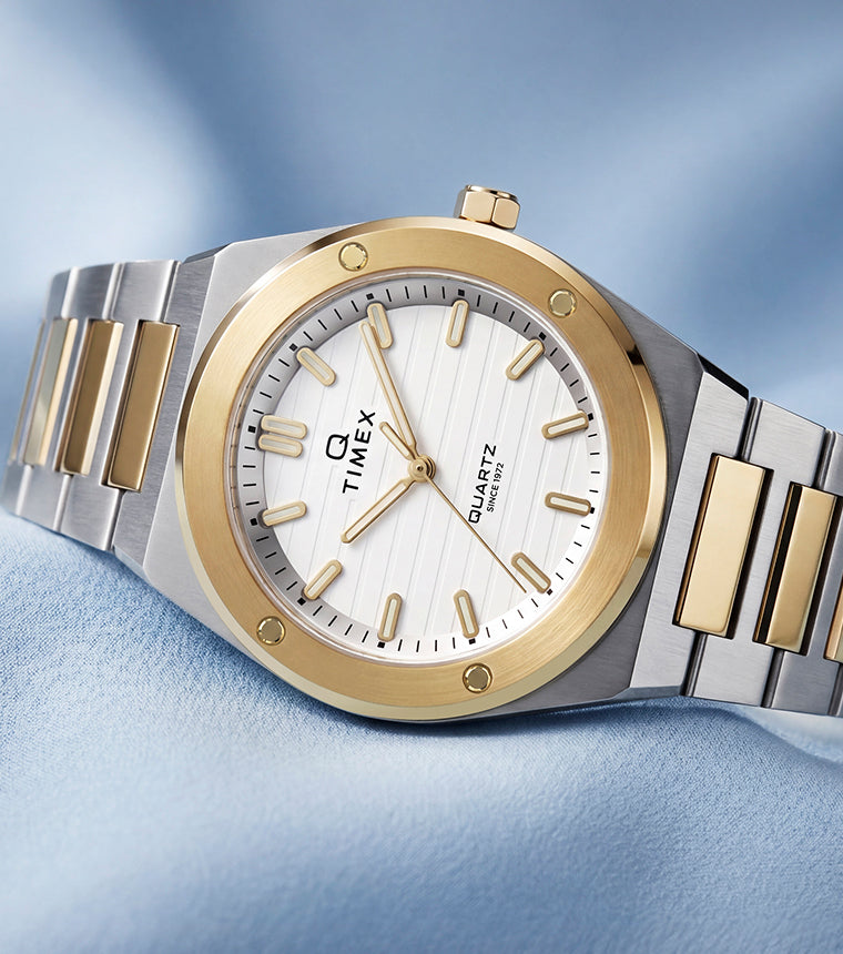 Q Timex Marbella two-tone stainless steel watch with a white dial and gold-tone bezel displayed on soft blue fabric.