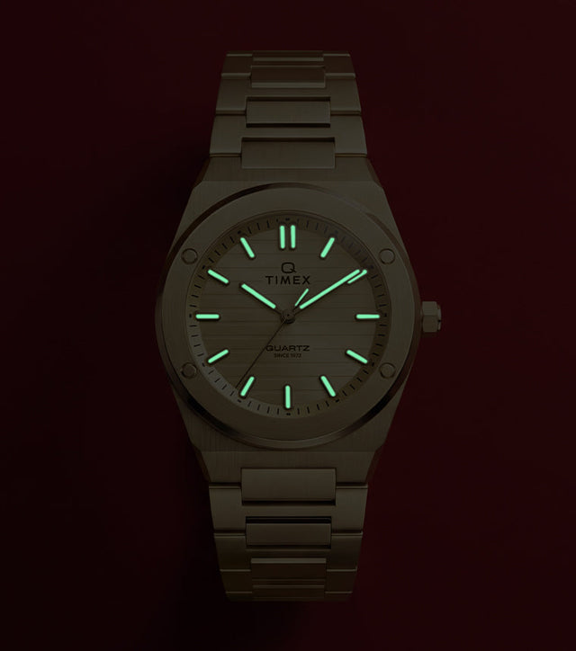 Q Timex Marbella gold-tone stainless steel watch with glowing Indiglo® hands and hour markers against a red background.