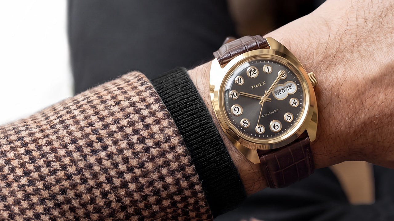 Timex® 1976 Rotary Phone Dial Reissue with a gold-tone case, black dial, and brown leather strap worn on the wrist with a patterned blazer.