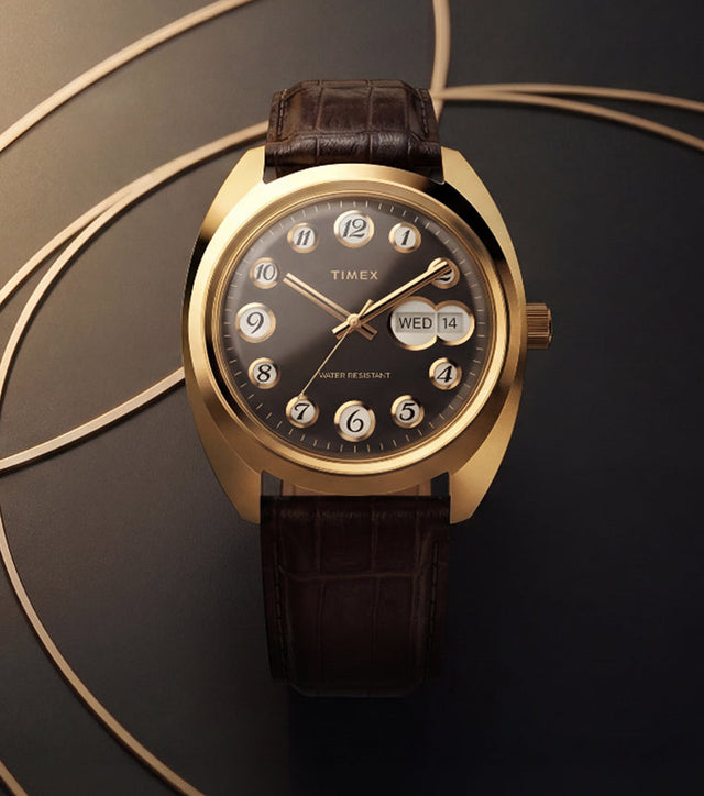Timex® 1976 Rotary Phone Dial Reissue watch with a gold-tone cushion case, black dial, rotating numeral hour markers, day-date display, and brown leather strap against a geometric gold-accent background.
