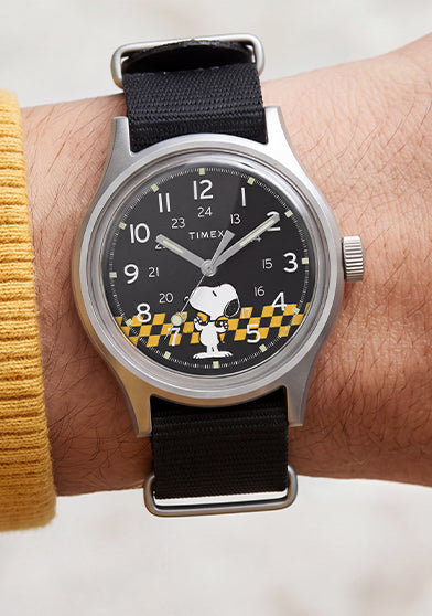 Peanuts® x Timex MK1® watch worn with a black fabric strap, featuring a black dial with Snoopy in sunglasses and yellow racing-inspired accents.