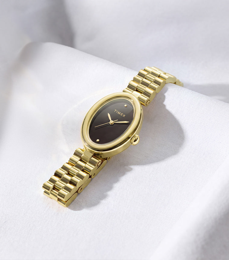 Gold-tone Timex Cavatina® Luxe watch with a dark oval dial and polished metal bracelet, displayed on softly draped white fabric.