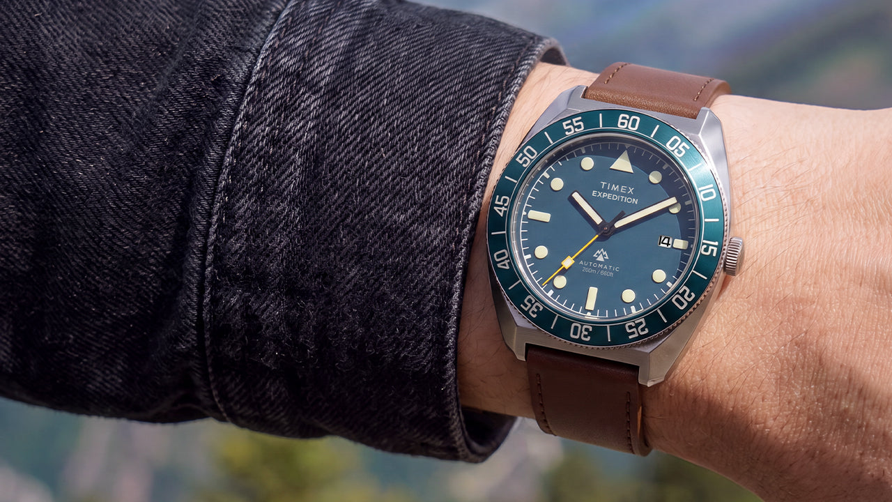 Timex Expedition Pioneer Automatic worn on the wrist with a dark jacket, featuring a blue dial, blue rotating bezel, and brown leather strap.