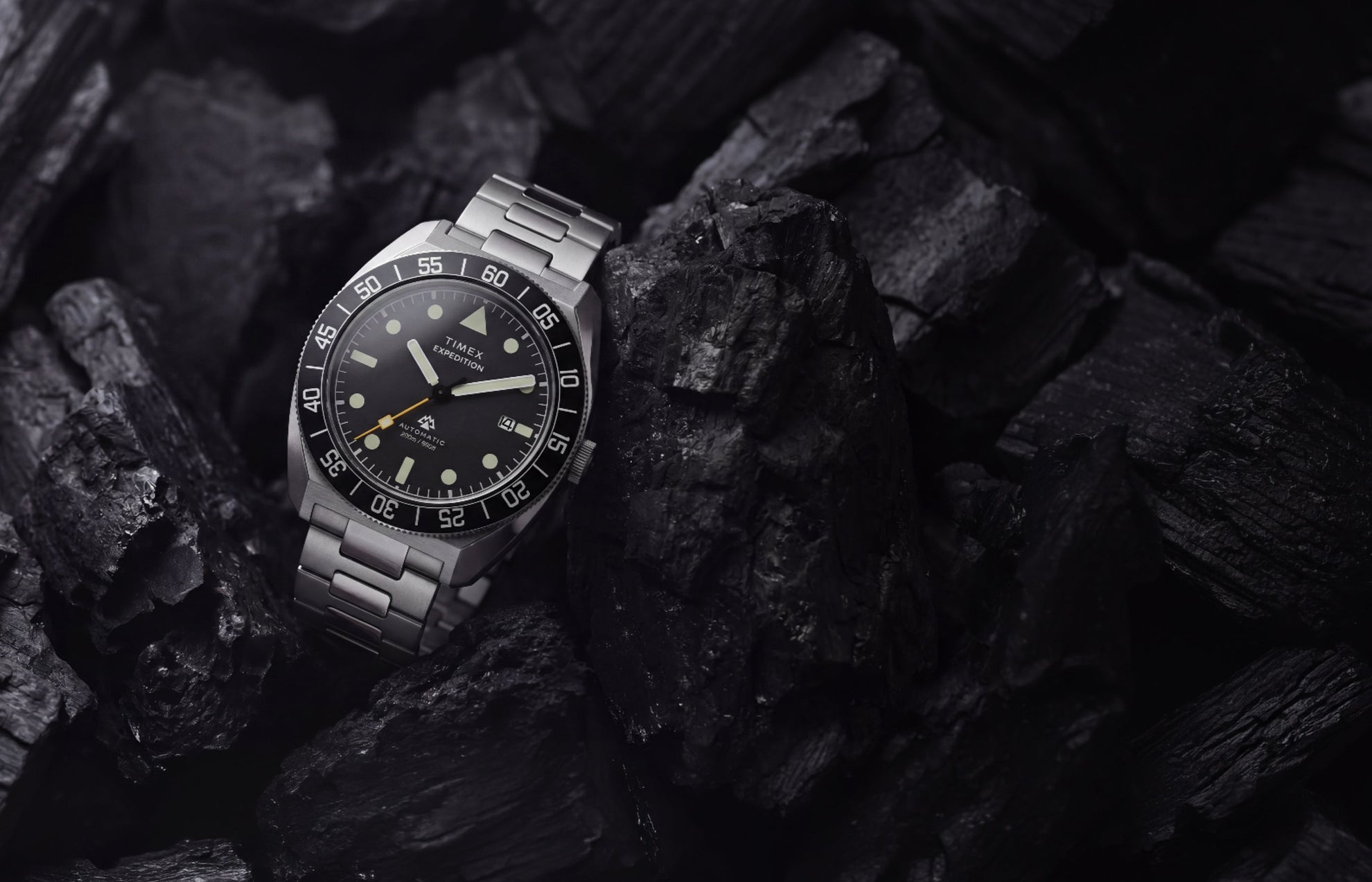 Timex Expedition Pioneer Automatic with a black dial, black rotating bezel, and stainless-steel bracelet displayed among dark charcoal rocks.