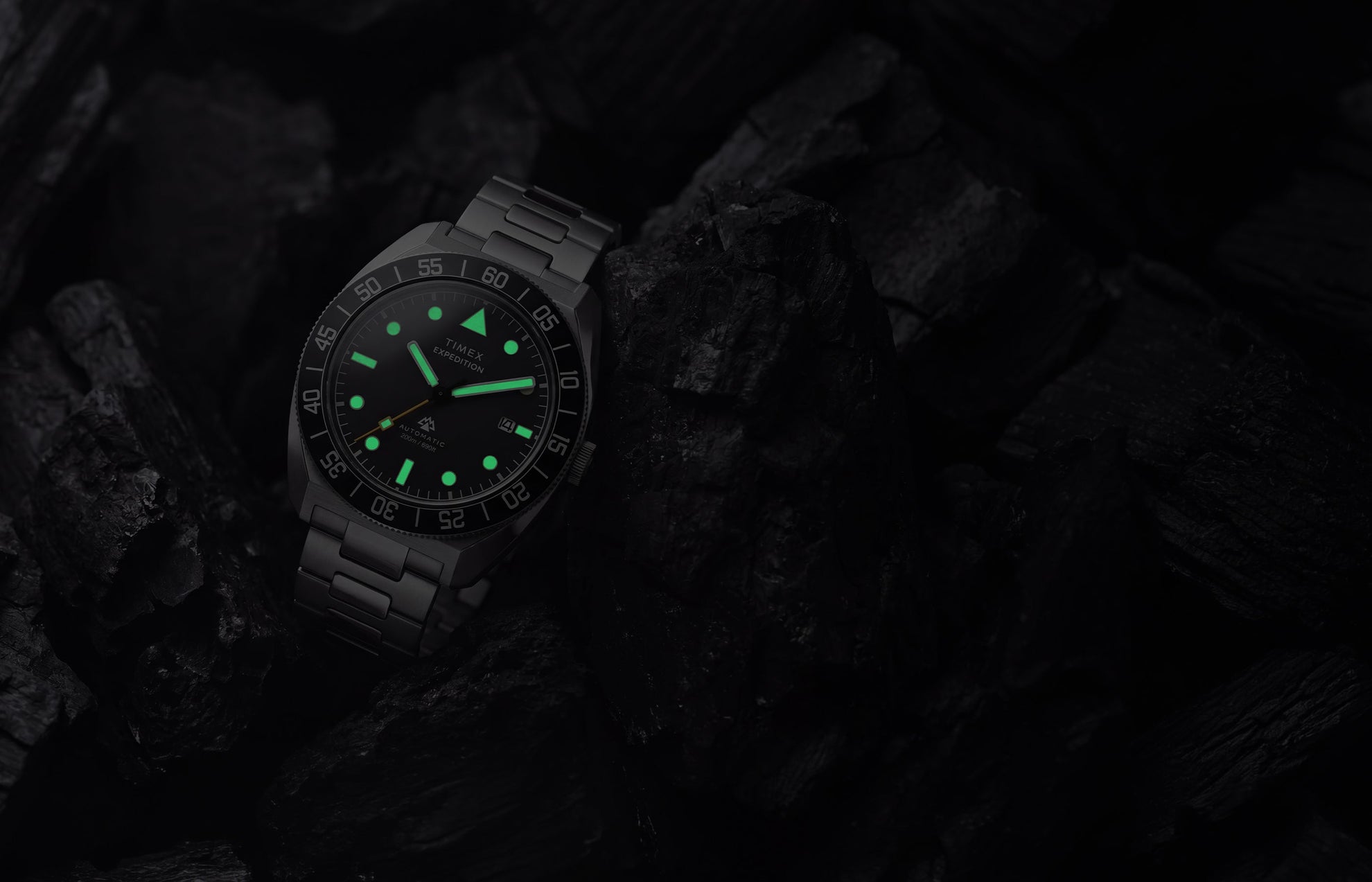 Timex Expedition Pioneer Automatic in low light with green luminous hands and hour markers glowing among dark charcoal rocks.