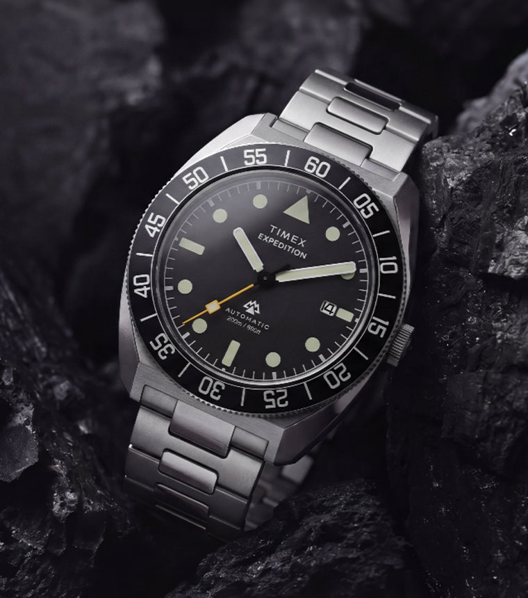 Timex Expedition Pioneer Automatic with a black dial, black rotating bezel, and stainless-steel bracelet displayed among dark charcoal rocks.