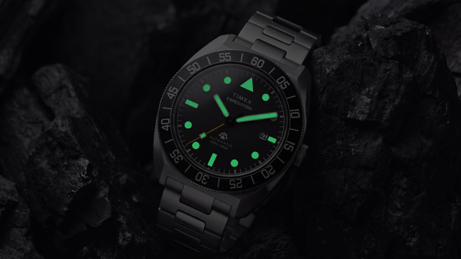 Timex Expedition Pioneer Automatic in low light with green luminous hands and hour markers glowing against dark charcoal rocks.