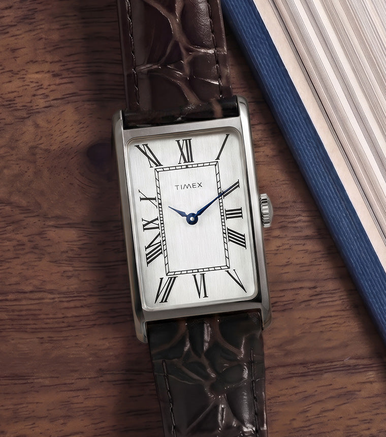 Timex 1976 Lexington Reissue Inspired watch with a rectangular silver-tone case, silver dial, blue hands, and brown crocodile-grain leather strap resting beside an open book on a wooden table.