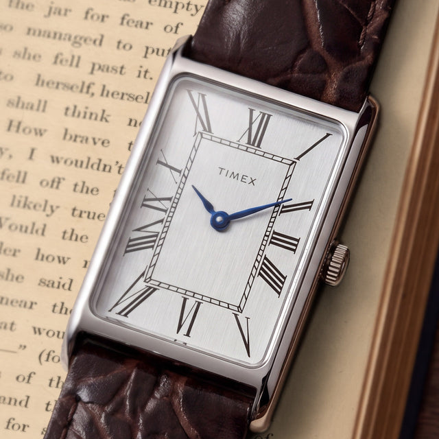 Close-up of the Timex 1976 Lexington Reissue Inspired watch featuring a silver sunray dial, black Roman numeral markers, blue hands, and a brown crocodile-grain leather strap.