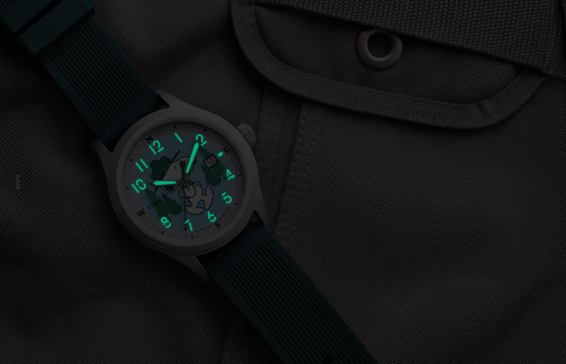 Peanuts® x Timex Expedition® Scout watch with illuminated green Indiglo® dial, highlighting the glowing Arabic numerals, hour markers, and hands against a dark gray tactical fabric background.