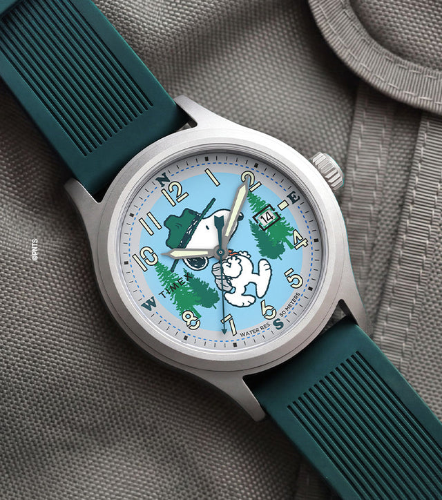 Peanuts® x Timex Expedition® Scout watch with a silver-tone case, light blue dial, and dark green silicone strap, featuring Snoopy hiking through a forest scene, luminous Arabic numerals, a date window, and displayed on a gray tactical fabric background.