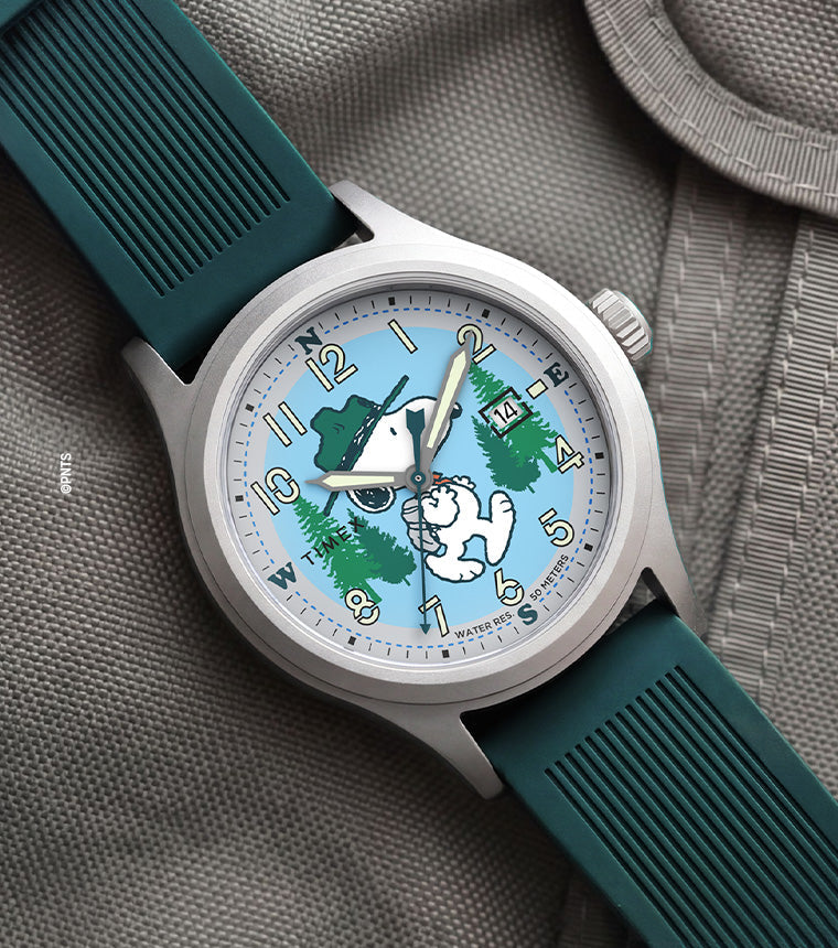 Peanuts® x Timex Expedition® Scout watch with a silver-tone case, light blue dial, and dark green silicone strap, featuring Snoopy hiking through a forest scene, luminous Arabic numerals, a date window, and displayed on a gray tactical fabric background.